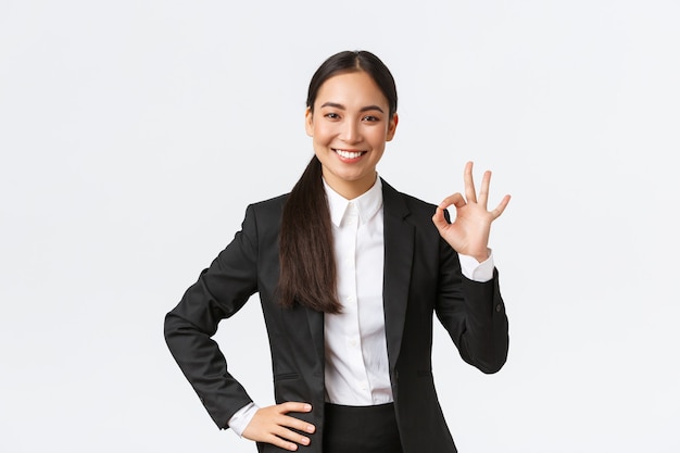 Professional female manager in black suit looking assertive, encourage everything okay, assure work done, showing okay gesture and smiling in approval, standing satisfied over white background