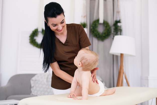 A professional female masseuse makes a massage to a little baby. Children's massage on the couch in a modern cozy room.