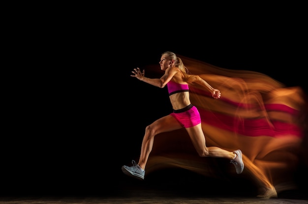 Professional female relay racer training isolated on black studio background in mixed light. Woman in sportsuit practicing in running. Healthy lifestyle, sport, workout, motion, action concept.