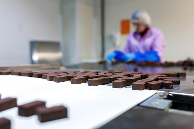 Professional female worker in uniform and protective gloves sorts chocolate candies on production line at factory