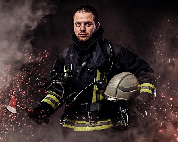 A professional firefighter dressed in uniform holding safety helmet in fire sparks and smoke over a dark background.