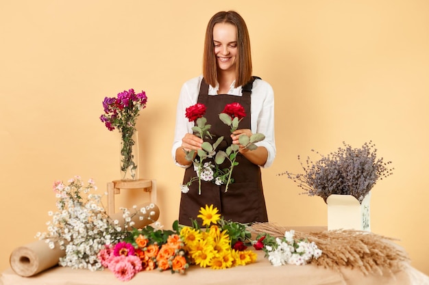 Professional florist shop Natureinspired floral design Hardworking joyful woman florist in brown apron making bouquet of fresh flowers holding roses in hands isolated over beige background