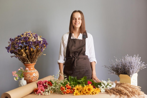 Professional florist workshop Fresh flower bouquet design Fragrant organic arrangements Smiling confident woman florist at workplace in flower shop standing isolated over gray background