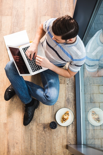 Professional freelancer. Top view of a positive nice delighted man sitting on the floor and holding a laptop while working as a freelancer