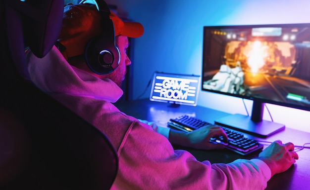 Professional Gamer Playing First-Person Shooter Online Video Game on His Powerful Personal Computer. Room and PC have Colorful Neon Led Lights. Young Man is Wearing a Cap at Home.