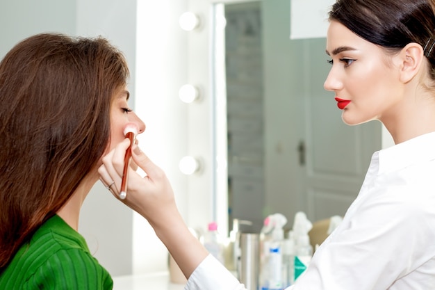 Professional make-up artist applying powder with blush on cheeks for young beautiful woman in beauty