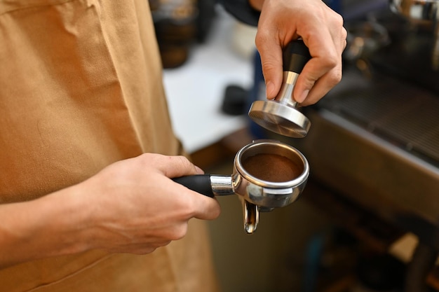 Professional male barista holding portafiler using coffee tamper to press coffee powder