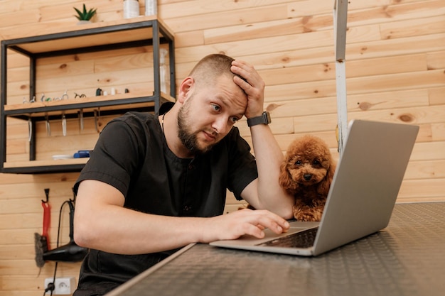 Professional male groomer working on laptop in her workplace in grooming salon near with poodle dog Animals Grooming concept