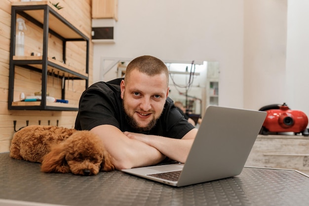 Professional male groomer working on laptop in her workplace in grooming salon near with poodle dog Animals Grooming concept