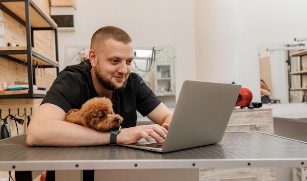 Professional male groomer working on laptop in her workplace in grooming salon near with poodle dog Animals Grooming concept