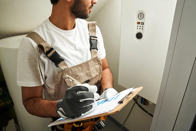 Photo professional male handyman in uniform is checking gas boiler at home and making notes in tablet