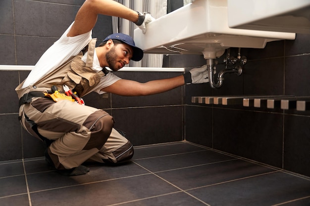 Professional male plumber in uniform is repairing a bathroom sink at home