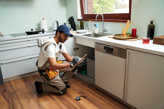 Photo professional male plumber in uniform sitting near kitchen sink and writing on clipboard in kitchen