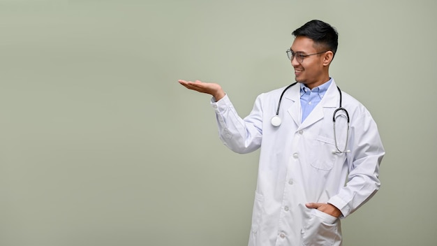 Professional millennial Asian male doctor in a white coat opens his palm looking aside