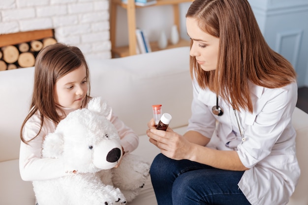 Professional pleasant doctor holding pills while giving tipps to little girl who is sitting with her fluffy toy