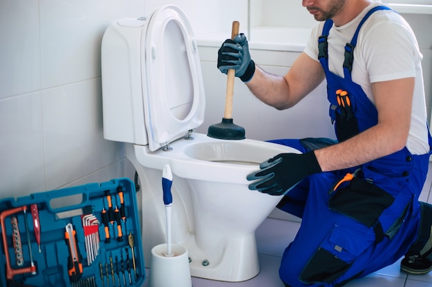 Professional plumber in uniform is repairing toilet bowl in the domestic bathroom with the help of tools