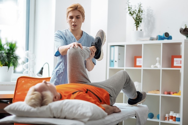 Professional rehabilitation. Nice blonde woman holding her patients leg while doing the rehabilitation exercise