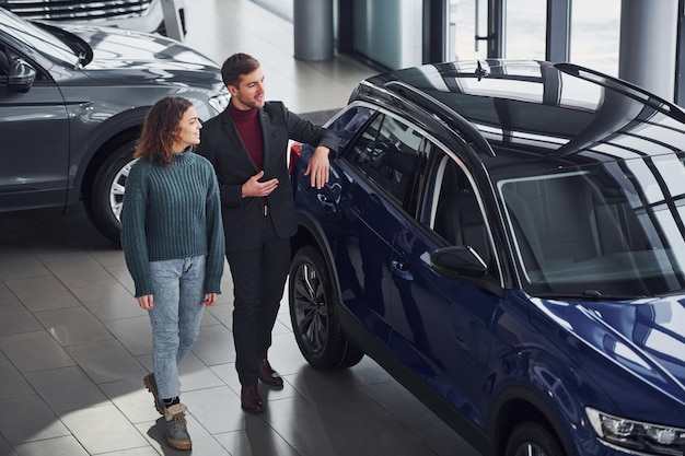 Professional salesman assisting young girl by choosing new modern automobile indoors.