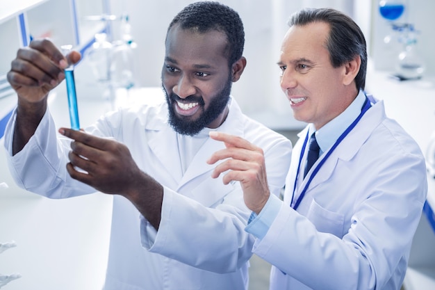 Professional scientists. Pleasant nice positive scientist standing behind his colleague and smiling while looking at the test tube