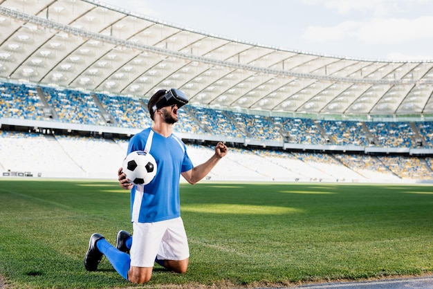 Photo professional soccer player in vr headset and blue and white uniform with ball standing on knees and