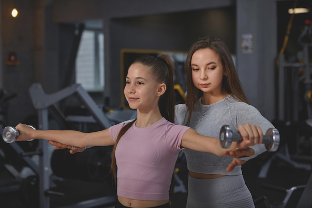 Professional sportswoman supervising teenage girl during gym workout doing dumbbells exercise