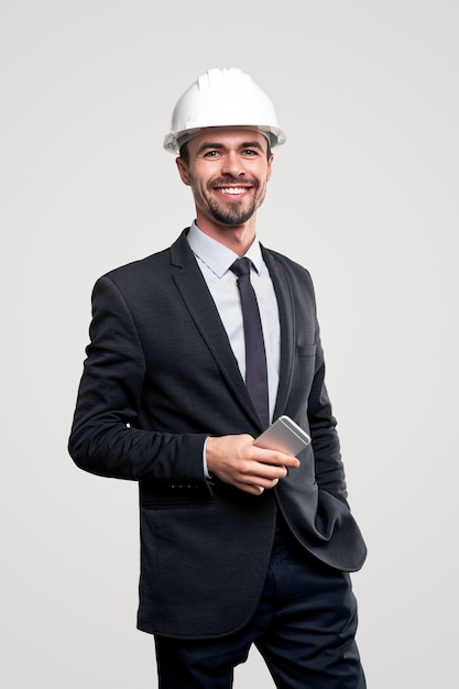 Professional successful male architect in elegant formal suit and hardhat with mobile phone in hand smiling happily and looking