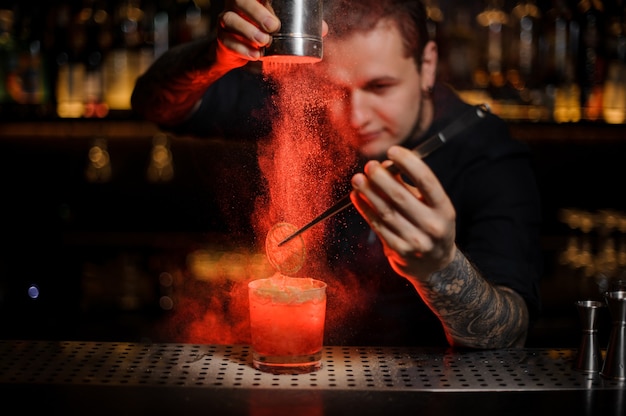 Professional tattooed bartender adding to an alcoholic cocktail in the glass a dried orange with tweezers and aromatic powder in the red light on the bar counter.