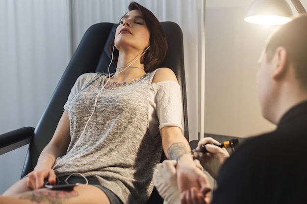 A professional tattooer artist doing picture on woman hand by machine while girl listening music. Tattoo art on body. Equipment for making black ink from a jartattoo art.