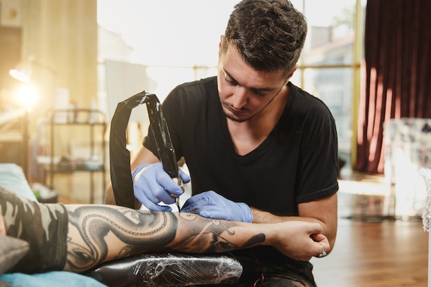 A professional tattooer artist doing tattoo on the arm of a young man by machine with black ink