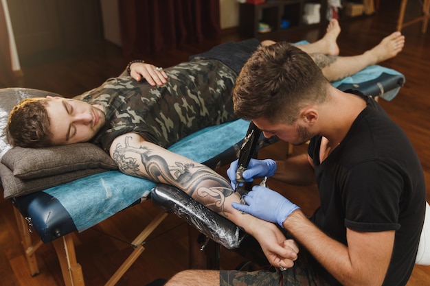 A professional tattooer artist doing tattoo on the arm of a young man by machine with black ink