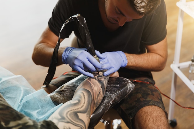 A professional tattooer artist doing tattoo on the arm of a young man by machine with black ink