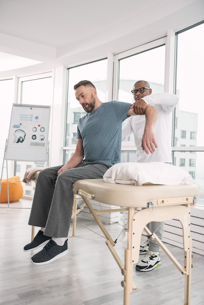 Professional therapy. Smart male therapist standing behind his patient while holding his arm