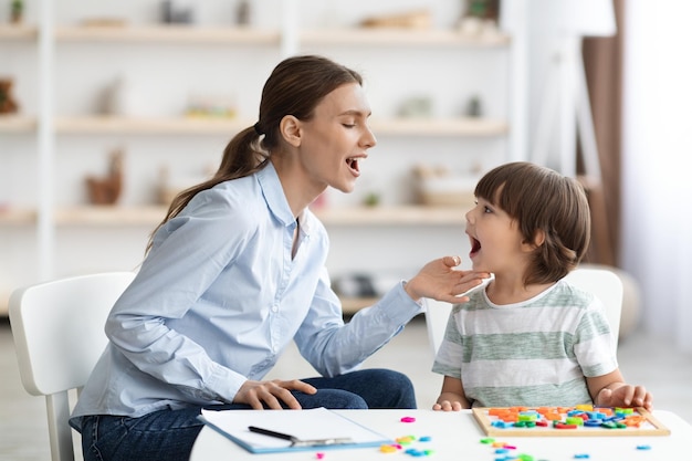 Professional woman speech therapist helping little boy to pronounce right sounds showing mouth