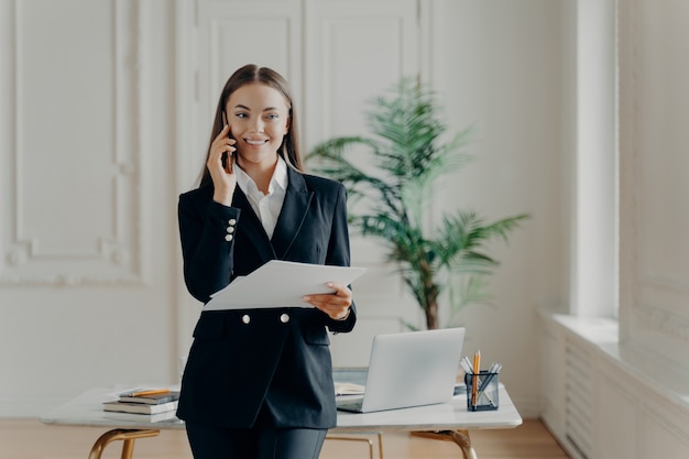 Professional young business lady in formal suit looking away while talking on phone and holding notes, business leader laughing when hearing good news, happy female office worker standing at office