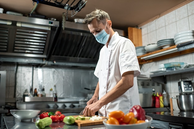 Photo professional young chef in uniform and protective mask chopping fresh zucchini and other vegetables while standing by large table