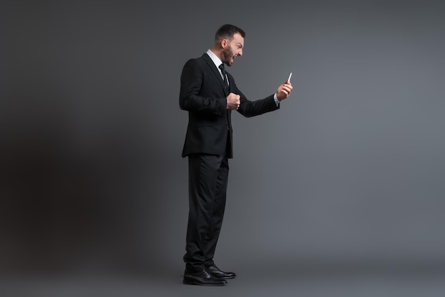 Profile of angry businessman in suit screaming while looking at smartphone on grey