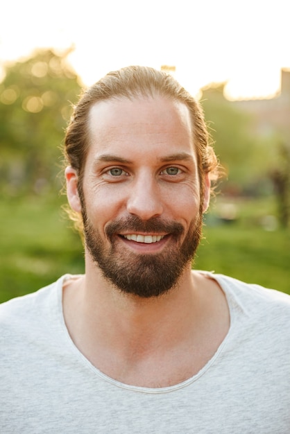 Profile closeup of handsome bearded man 30s with tied hair in white t-shirt smiling, while resting in green park