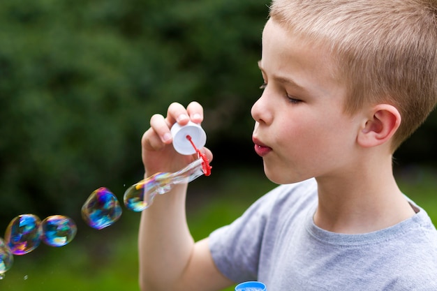 Profile of cute handsome small blond child boy with funny serious expression blowing colorful transparent soap bubbles outdoors on blurred green summer background. Joy of careless childhood concept.