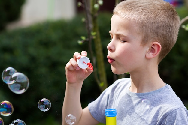 Profile of cute handsome small blond child boy with funny serious expression blowing colorful transparent soap bubbles outdoors. Joy of careless childhood concept.