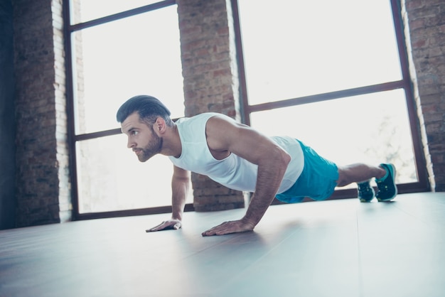 Profile photo of macho sportsman guy do push ups plank hands leaning floor determined trainer sportswear tank-top shorts sneakers training house  windows indoors