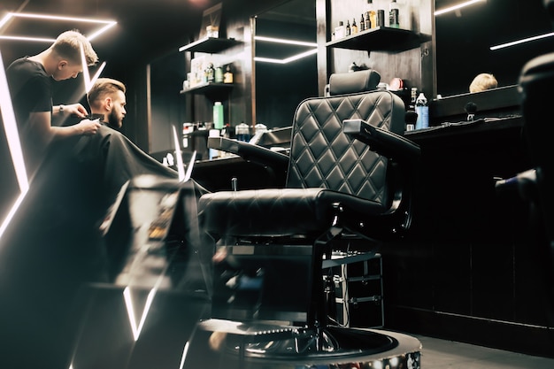 Profile photo of a young barber trimming his customerÃ¢ÂÂs hair with an electric shaver and a comb in a barbershop.