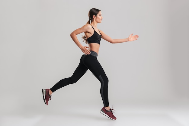 Profile picture of energetic caucasian female in sportswear running, isolated along gray wall