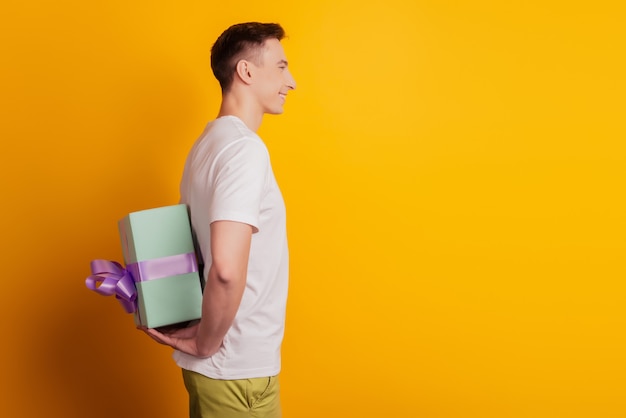 Profile portrait of cheerful nice guy hold persent package behind back make surprise on yellow background