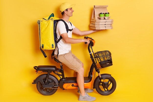 Profile portrait of courier man wearing cap and tshirt driving electric bike motorbike handing pizza in cardboard and takeaway coffee isolated on yellow background