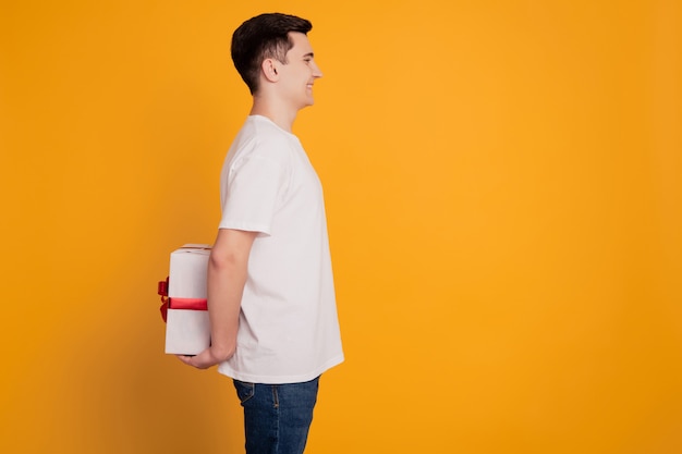 Profile portrait of funny guy hold gift package behind back look empty space make surprise on yellow background