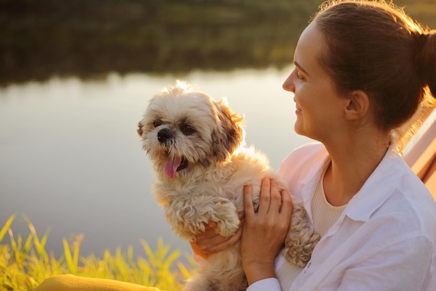 Profile portrait of smiling positive dark haired young adult woman wearing white shirt and her Pekingese dog sitting at sunset near the river vacation in nature