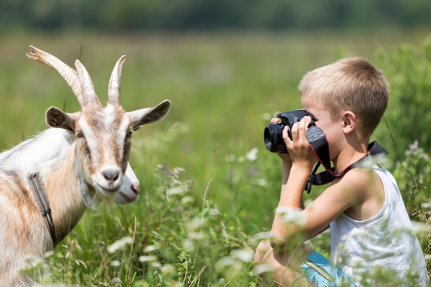 Profile portrait of young blond cute handsome child boy taking picture of funny curious goat.