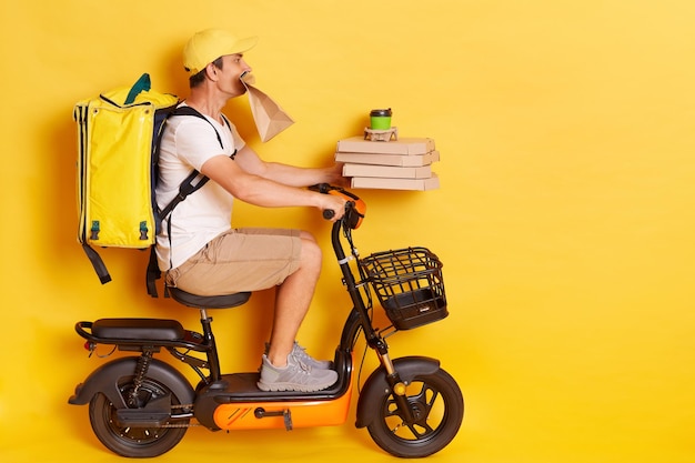 Profile portrait of young man with backpack wearing white t shirt riding escooter with pizza boxes and coffee holding paper parcel in teeth delivering lots orders isolated over yellow background