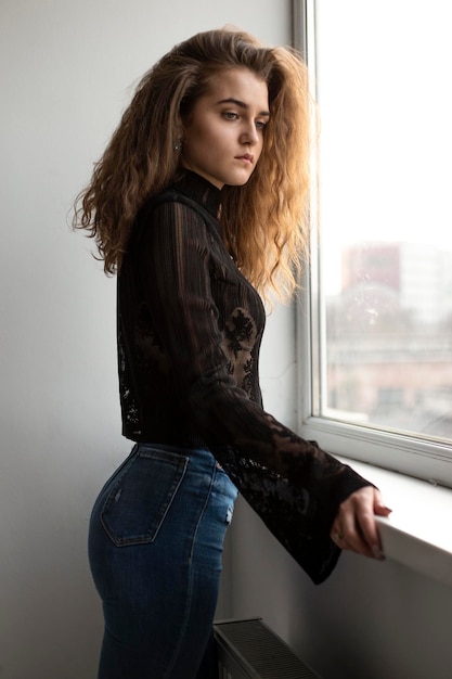 Profile portrait of a young woman with curly brown hair dressed in a black suitcase and jeans posing near window