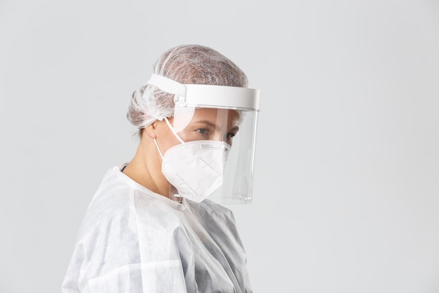 Profile of serious-looking female doctor in personal protective equipment, face shield and respirator listening to patient, provide checkup.